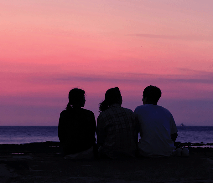 Silhouette of three friends sitting on a rocky coastline at sunset.