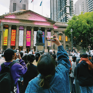 Woman takes a photo on a phone of a large crowd gathered in front of a neoclassical building.