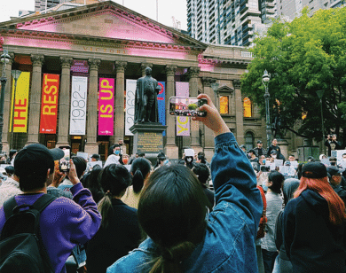 Woman takes a photo on a phone of a large crowd gathered in front of a neoclassical building.