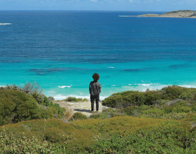 A person stands on rocks close to the blue ocean.